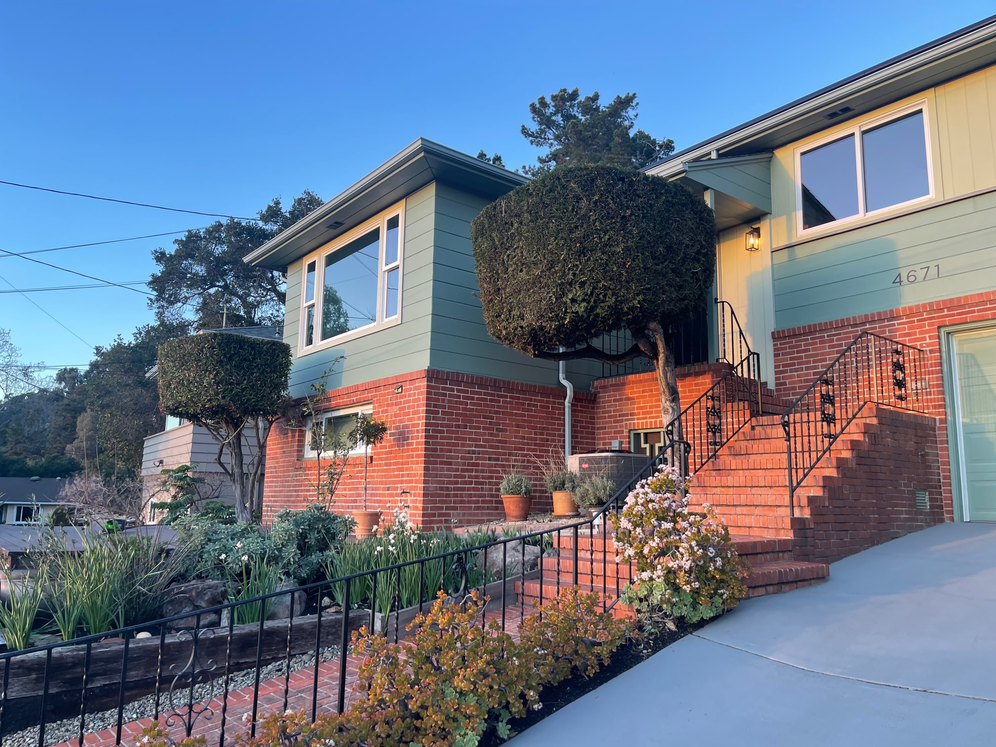 Two-story green and brick house with brick stairs, black railings, and manicured square trees.