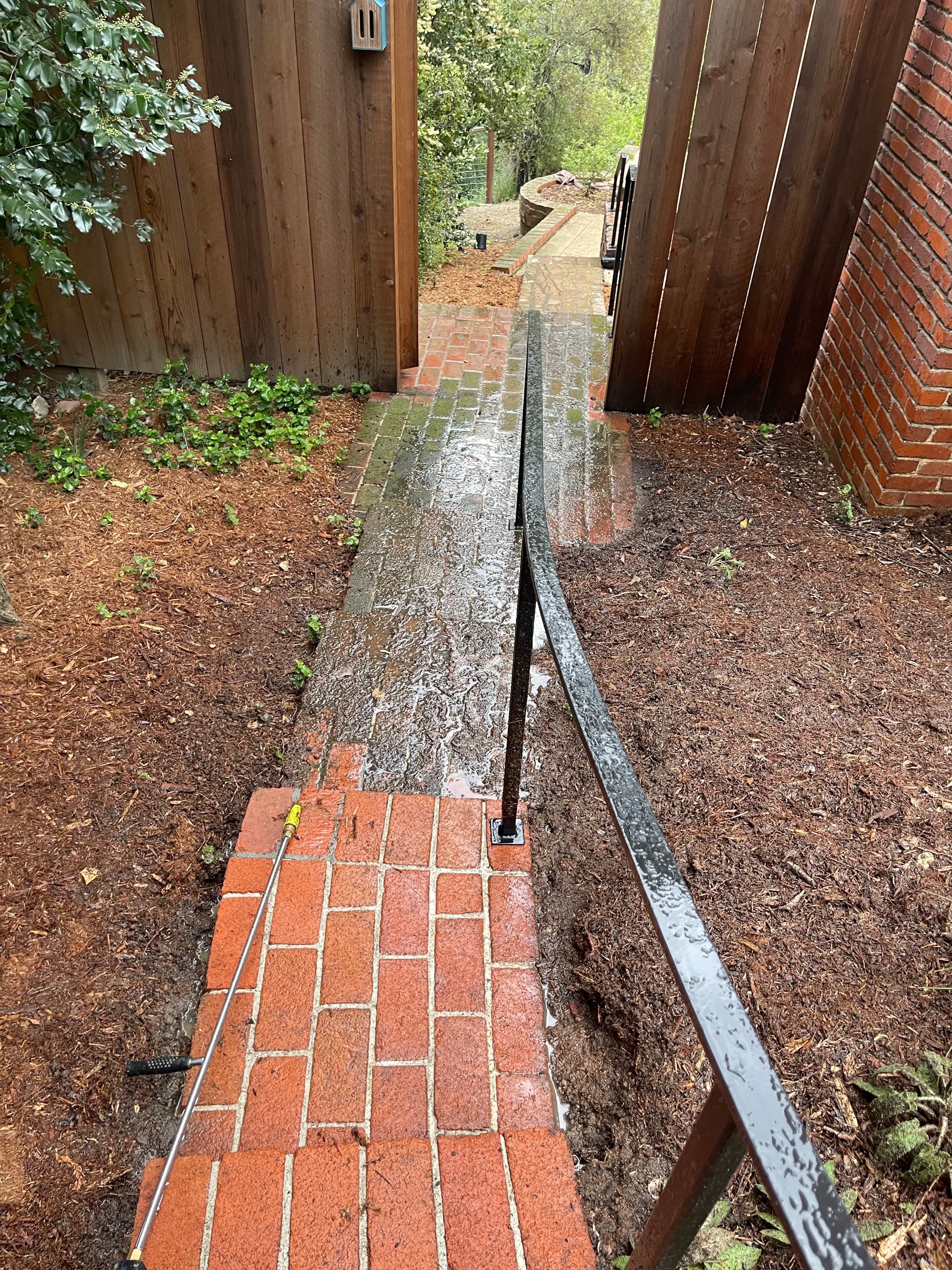 Pressure washer wand cleaning a mossy brick walkway, revealing bright red bricks beneath the grime.
