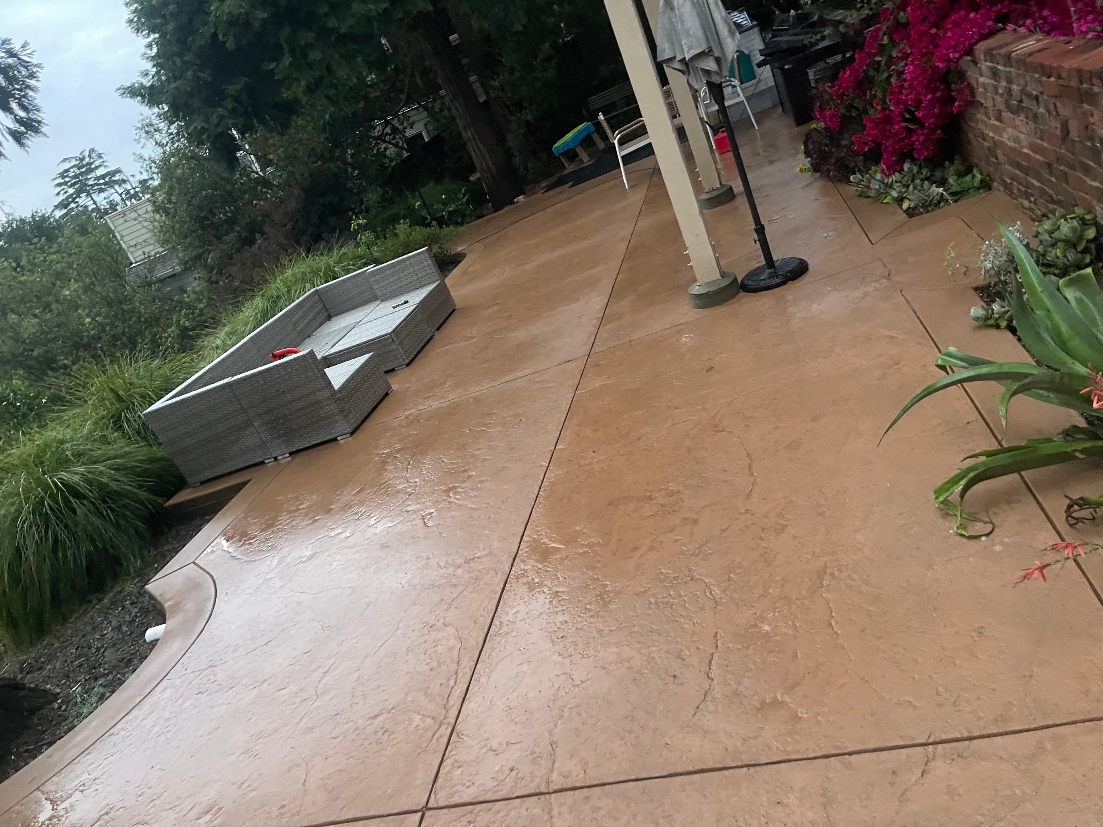 Tan stamped concrete patio with grey wicker sofa, lush greenery, and vibrant pink flowers.